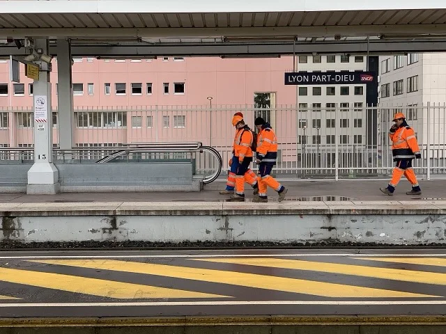 Láthatósági ruházatot viselő dolgozók vasúti pályaudvaron, munkavédelmi felügyelet mellett